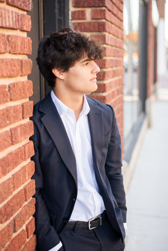 Senior guy wearing a dark suit and white dress shirt leans against a brick wall looking to side. senior pictures clothes ideas for guys