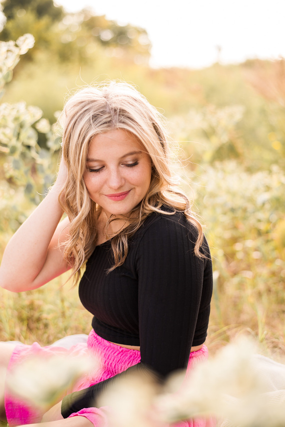 high school senior girl with long blond hair in a black top and pink skirt sits among wildflowers in a field in central Oklahoma smiling softly with one hand in her hair