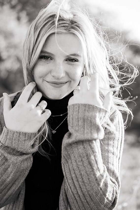 black and white portrait of a high school senior girl with long hair blowing in the wind as she holds it back softly with her hands and smiles at the camera in a field near Oklahoma City