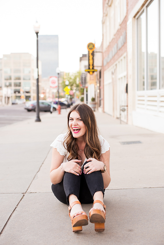 pretty senior girl with long brown hair sits cutely hugging her knees and smiling joyfully on a sidewalk in Midtown OKC with Broadway Street buildings behind her