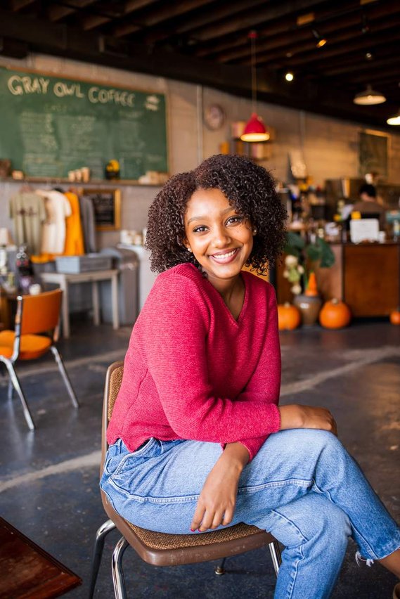 Black high school senior girl in blue jeans and a pink sweater sits and smiles with legs crossed in a brown chair inside Gray Owl coffee shop in Norman, Oklahoma