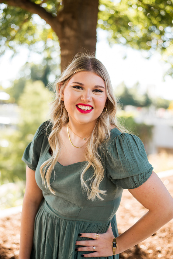portrait of a high school senior girl with long blond hair and bright pink lips in a green dress standing with her hand on her hip and smiling