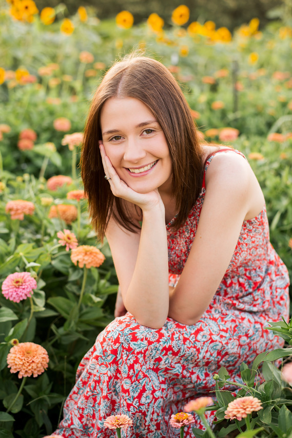 High school senior girl sits with her chin in her hand among zinnias and sunflowers at a flower farm in Oklahoma
