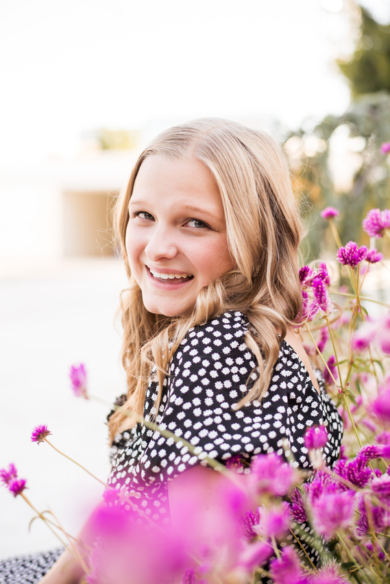 close up portrait of a blond high school senior girl wearing a black and white dress sitting among pink flowers and smiling over her shoulder