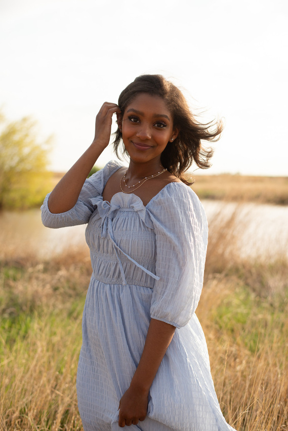 straight out of camera image of a high school senior girl wearing a blue dress standing in a field with a pond behind her holding back her hair in Tuttle Oklahoma