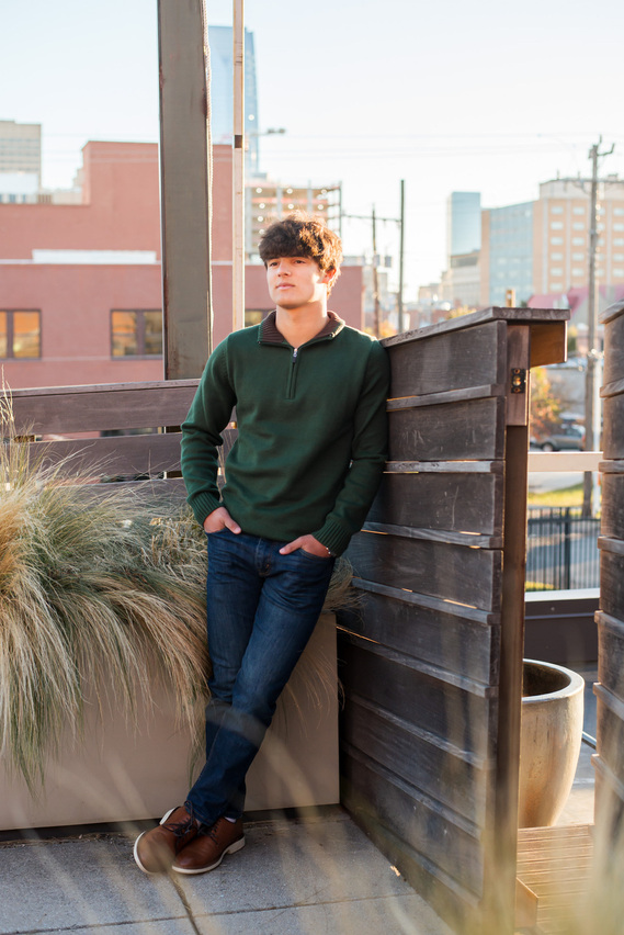 senior boy wearing a dark green sweater and dark jeans with dress shoes leans against a wood fence with fall grass beside him on a rooftop with the OKC downtown skyline behind him in the distance