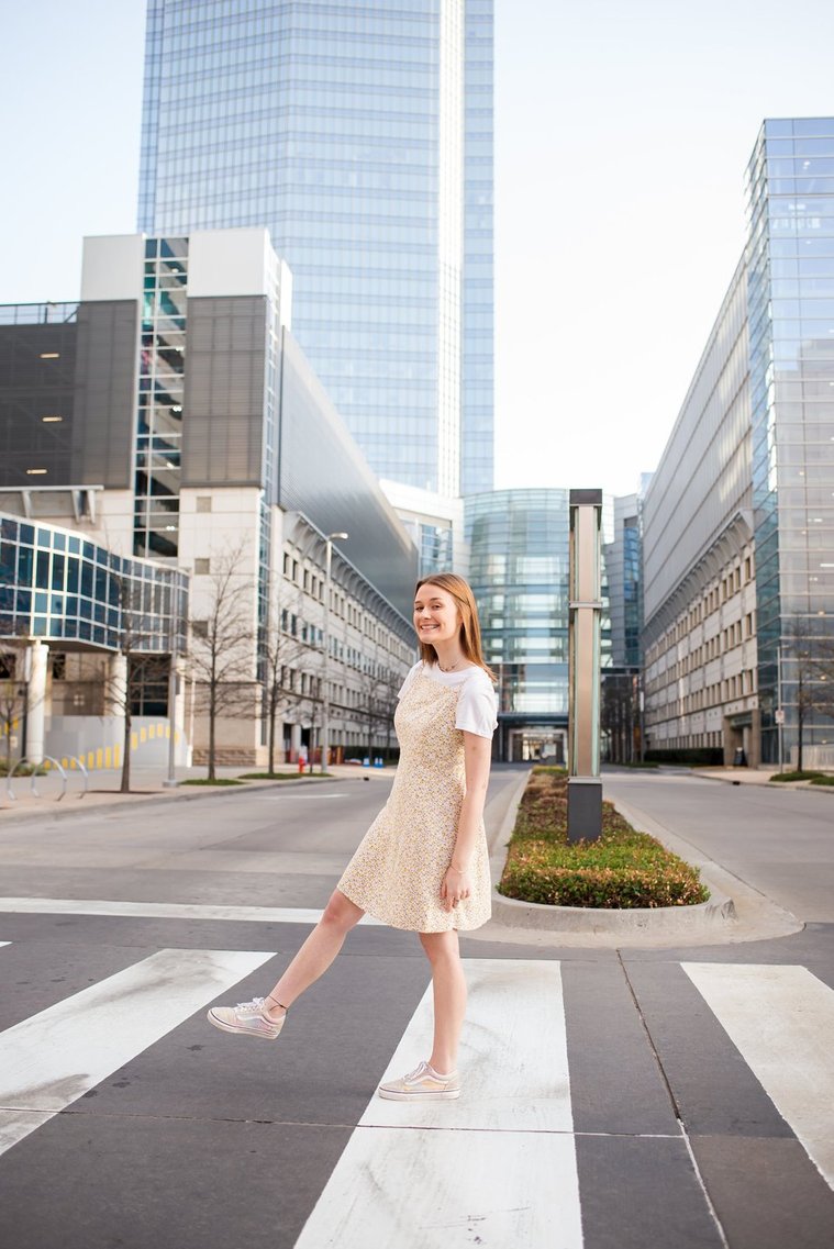 high school senior girl walks across a crosswalk kicking her leg up and smiling with the Devon tower in downtown Oklahoma City in the background