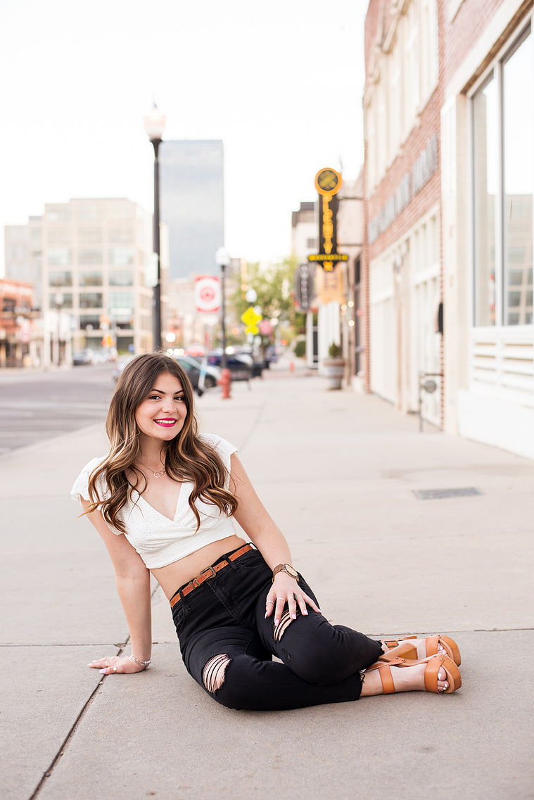senior photo of a beautiful girl long brown hair wearing a white top and black jeans sitting on her hip with legs folded and smiling on a sidewalk with midtown OKC behind her