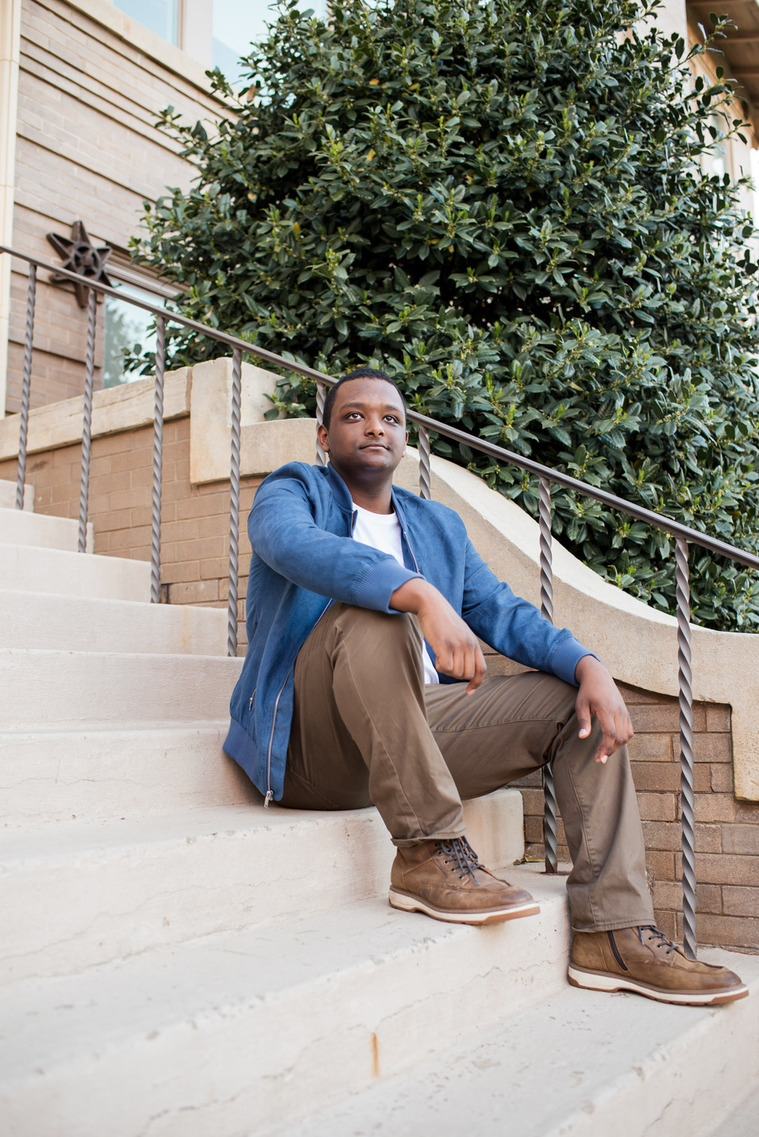 senior guy wearing a blue jacket and brown pants sits on steps with arms draped on legs looking off at OU campus in Norman, Oklahoma