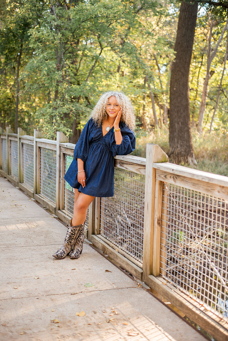 senior girl with curly blond hair wearing a blue denim dress and cowboy boots leans against a wooden footbridge over a small creek smiling softly at Martin Nature Park in Oklahoma City