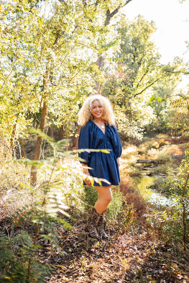 pretty senior girl with blond curly hair smiles and stands in the trees with a creek behind her at Martin Nature Park in OKC