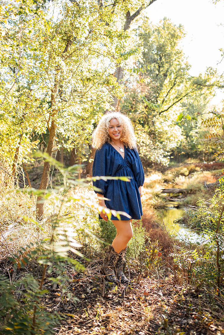 pretty senior girl with blond curly hair smiles and stands in the trees with a creek behind her at Martin Nature Park in OKC