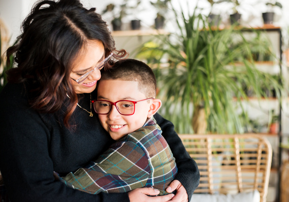 a mom and son hug and smile in front of plants and a wicker chair at a plant shop in Oklahoma City, Oklahoma