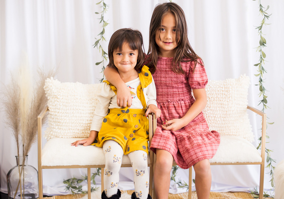 two little girl sisters sit on white chairs and hug each other and smile with a white background around them