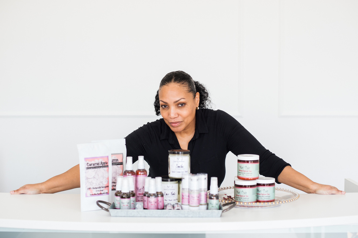 branding photo of a small business owner woman of color standing in a white room at a counter with her organic bath and body products in front of her