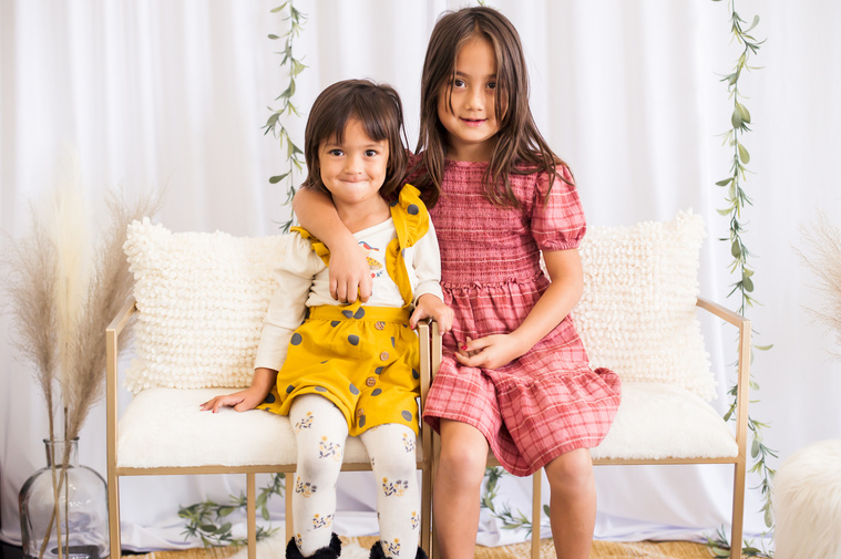 two little girls sit on white chairs and hug each other in front of a white curtain and green ivy in a studio in Oklahoma City