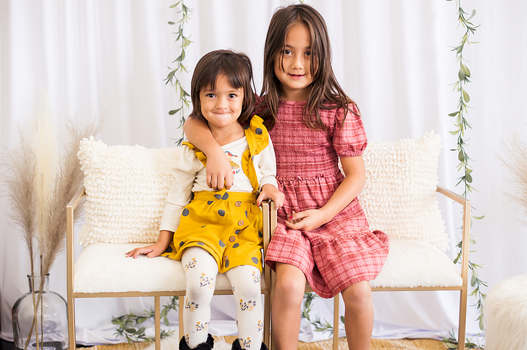 two little girls sit on white chairs and hug each other in front of a white curtain and green ivy in a studio in Oklahoma City