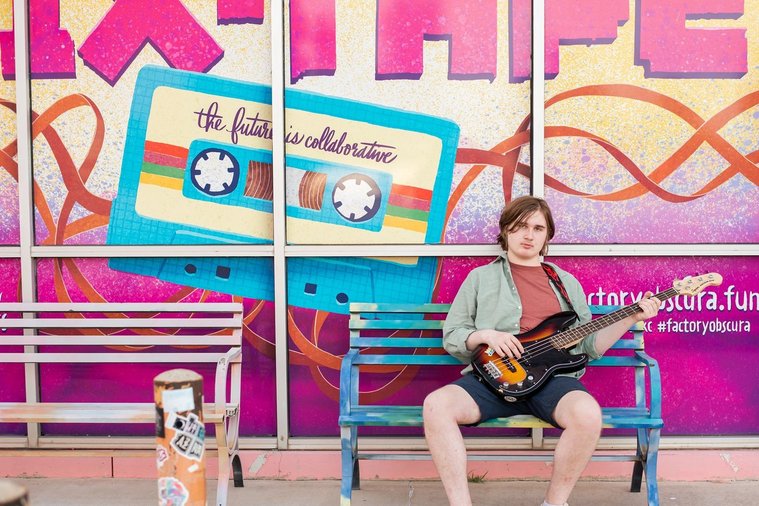 senior boy wearing a button down shirt and blue shorts sits on a bench holding his bass guitar and smiling in front of a wall mural in midtown Oklahoma City