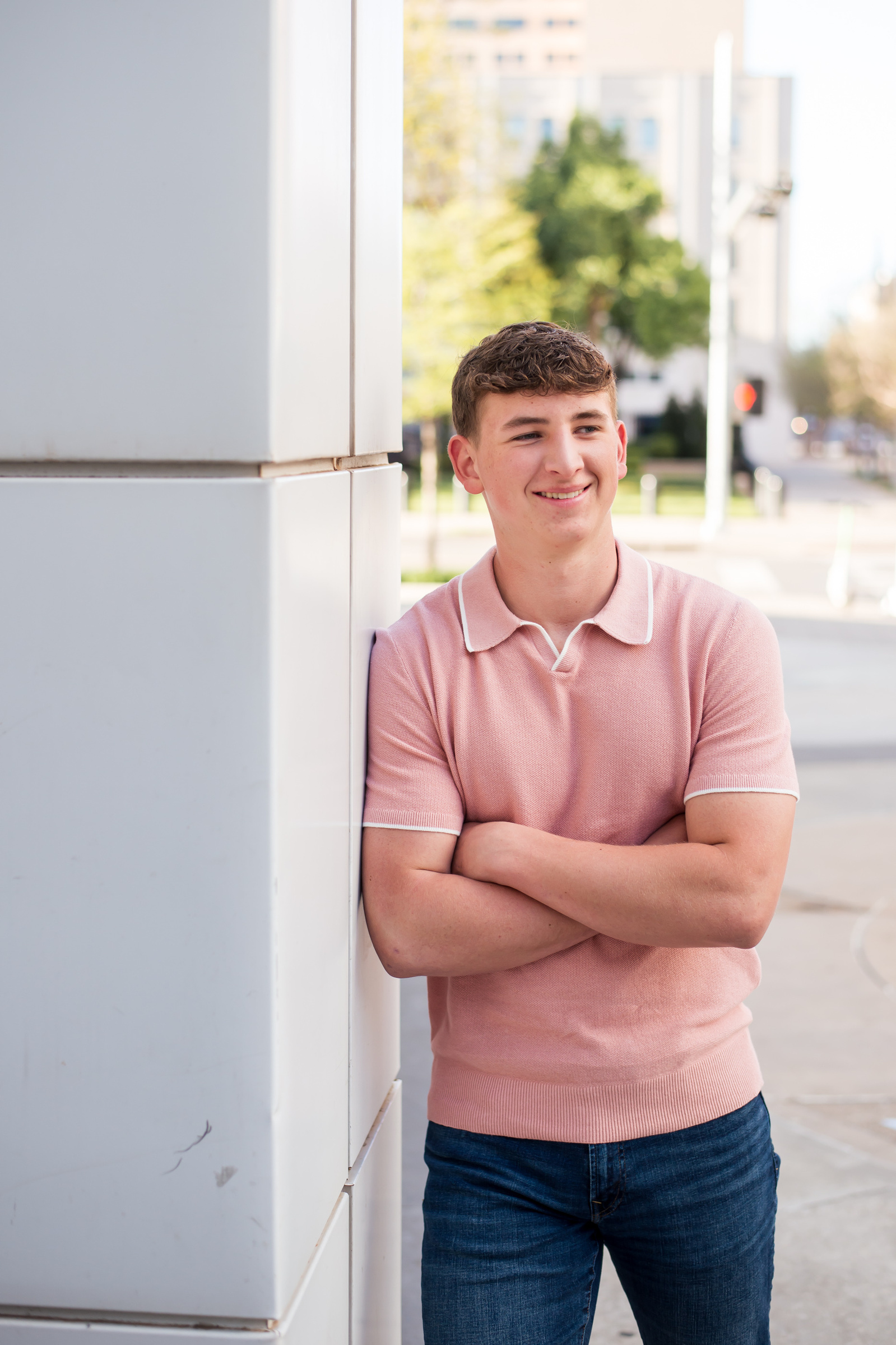 white high school senior boy wearing a peach polo shirt leans against a white pillar with arms crossed and smiling to the side in downtown Oklahoma City