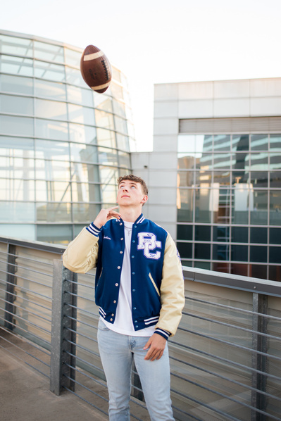 senior boy in a letterman jacket throws a football in the air on the top floor of a parking garage next to Devon Tower in OKC