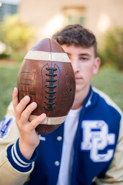 senior boy wearing a letterman jacket holds a football out covering half his face in an artistic senior portrait taken in downtown OKC