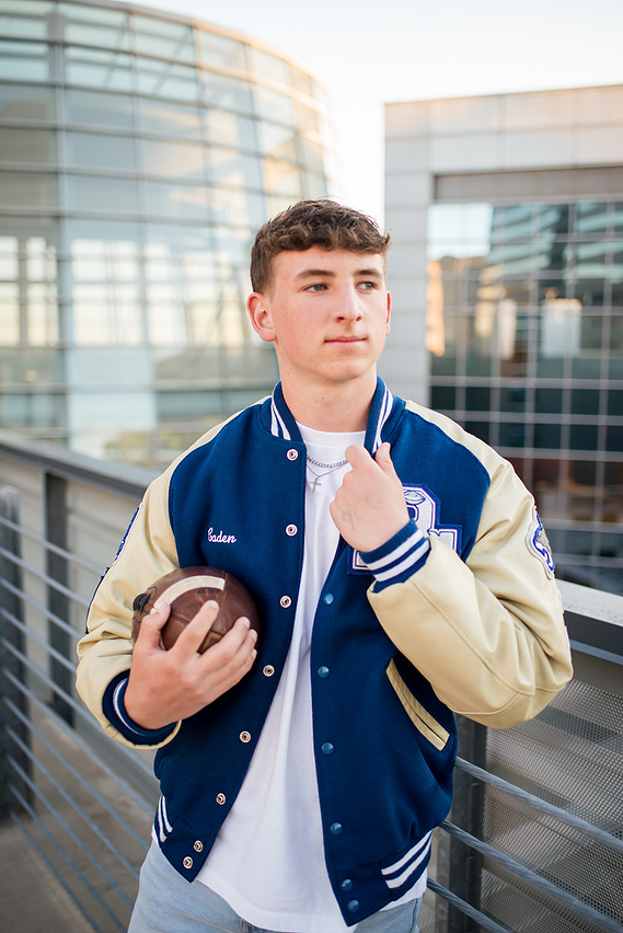 senior boy wearing his letterman's jacket holding a football poses seriously as he stands on the rooftop of the Devon parking garage in Oklahoma City with buildings around him
