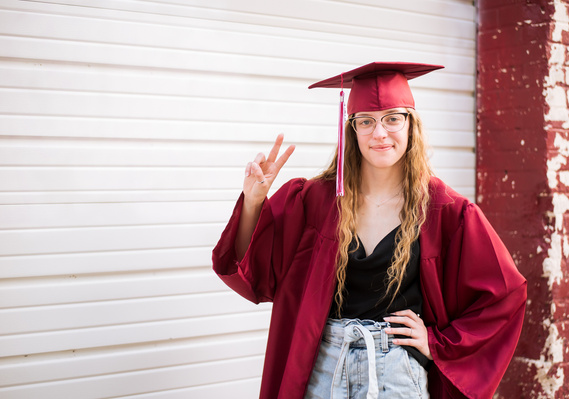 High school senior girl poses in her red cap and gown with a peace sign in front of a white rustic garage door and smiles at here senior photo session in Oklahoma City, OK