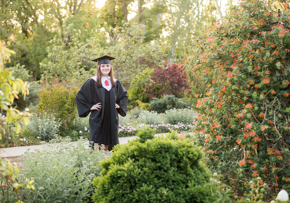 High school senior girl stands in her cap and gown in the middle of flowers and greenery at Will Rogers Gardens in Oklahoma  City, Oklahoma