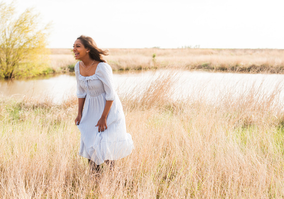 High school senior girl stands in a field of golden grass with a pond and laughs at her senior portrait session in central Oklahoma