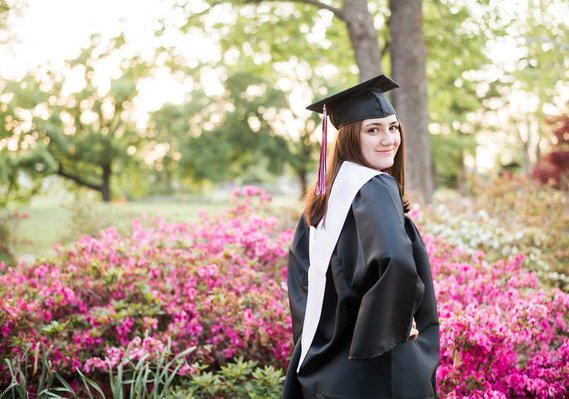 High school senior girl poses in her cap and gown in front of pink flowers at Will Rogers Gardens in Oklahoma City