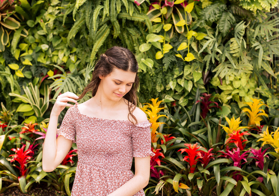 Senior girl wearing a red and white dress sits in front of a tropical plant wall and looks softly down her shoulder while holding end of her hair at Crystal Bridge Conservatory at Myriad Gardens in Oklahoma City