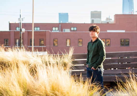 senior boy wearing a green sweater and jeans stands on the rooftop of Coffee Slingers in Automobile Alley in Oklahoma City surrounded by golden grass and the downtown skyline behind him