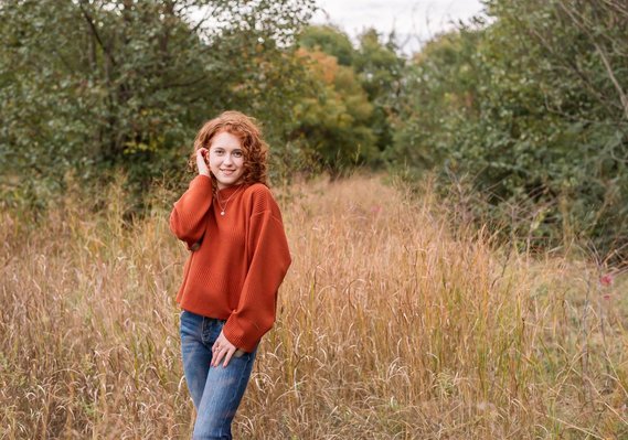 Senior girl with red hair wearing an orange sweater and jeans stands cutely and smiles in a field with gold grass and fall trees behind her at Sutton Park in Norman, Oklahoma