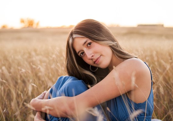 senior girl wearing a blue dress sits among tall golden grass hugging her knees to her chin and smiling softly in the fall in a field in Tuttle Oklahoma