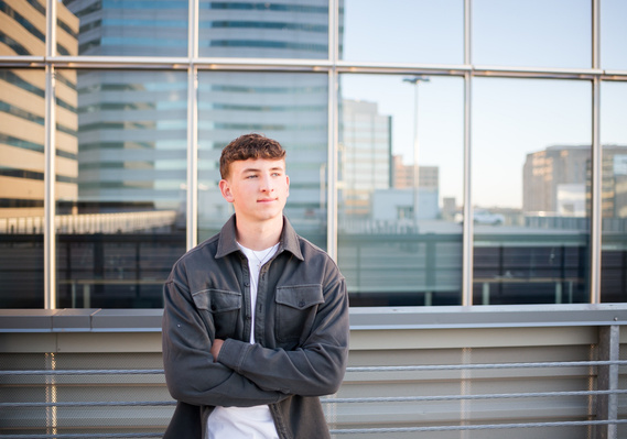 senior boy with arms crossed and smiling off to side stands on a parking garage rooftop in Oklahoma City with other buildings reflecting behind him