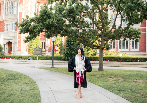 College senior girl wearing black gown and graduation stole stands and poses on a sidewalk on OU campus with Evans Hall behind her