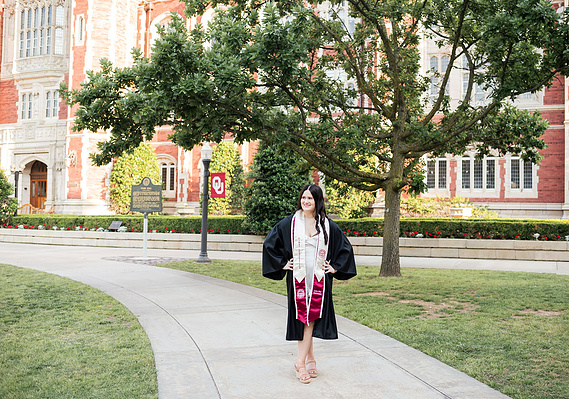 College senior girl wearing black gown and graduation stole stands and poses on a sidewalk on OU campus with Evans Hall behind her