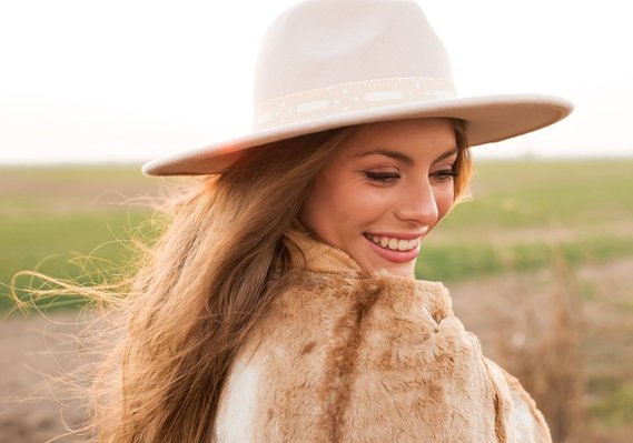 beautiful senior girl wearing a western hat and wrapped in a brown blanket smiles over shoulder standing in front of a field at sunset near Oklahoma City