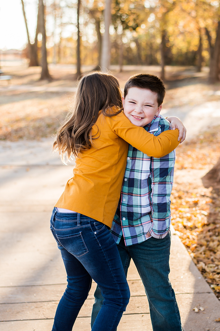 a young girl is hugging her brother tight while he smiles but isn't loving it at a family photo session in the fall at a park in Moore, Oklahoma
