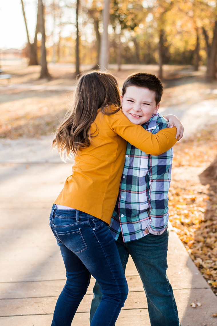 a young girl is hugging her brother tight while he smiles but isn't loving it at a family photo session in the fall at a park in Moore, Oklahoma