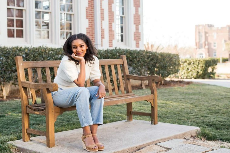 African-American teenage girl sits on a bench on OU campus and smiles at her cap and gown senior photo shoot in Norman Oklahoma