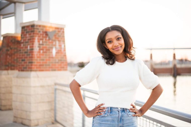 African-American teenage girl stands with her hands on her hips and smiles at Legacy Park at her cap and gown senior photo shoot in Norman Oklahoma