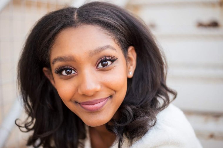 African-American teenage girl sits on white steps in a white sweater and smiles at her cap and gown senior photo shoot in Norman Oklahoma