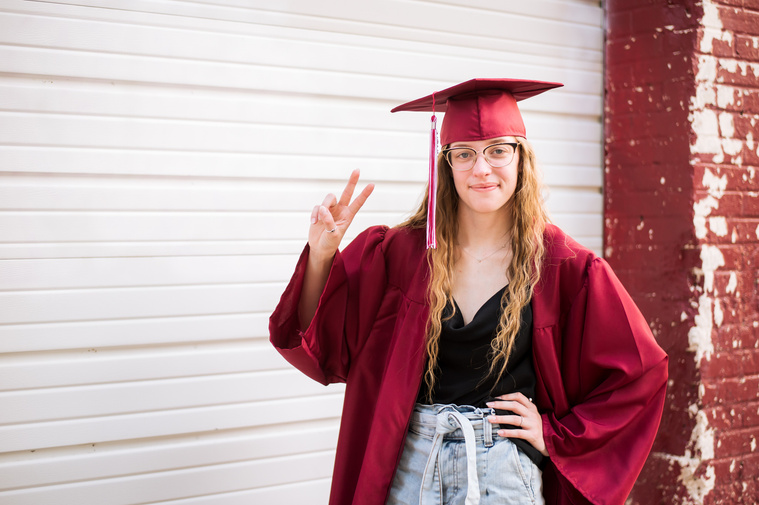 high school senior girl with long blond hair wearing her red cap and graduation gown poses with a peace sign in front of a rustic garage door in midtown Oklahoma City
