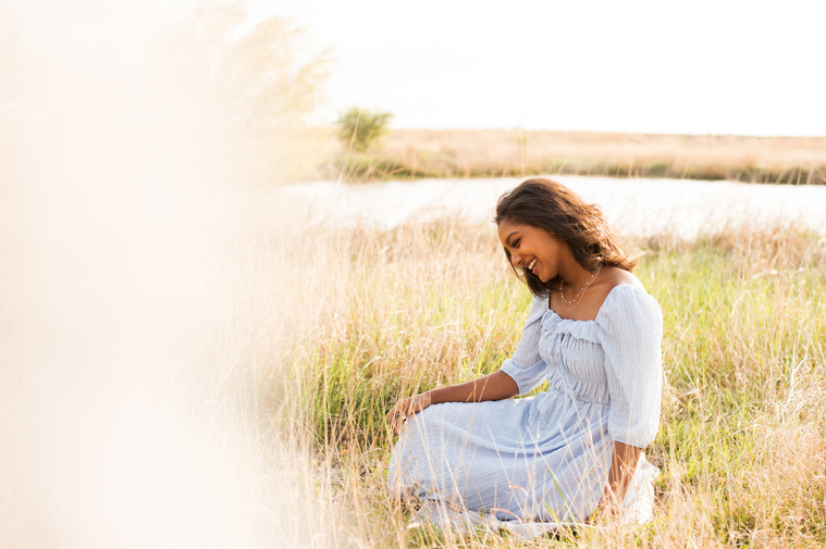black senior girl wearing a light blue dress sits among tall grass in a field near Oklahoma City with a pond behind her and is smiling down happily with a blurry cream foreground to the side of her