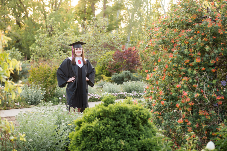 High school senior girl stands in her cap and gown with greenery and flowers around her at Will Rogers Gardens in Oklahoma City for a senior photo session