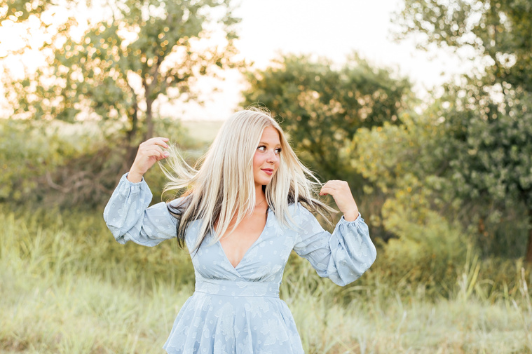 Artsy image of a High school senior girl looking to the side and holding each side of her hair with a hand in a blue dress  in a field in central Oklahoma for her senior photos