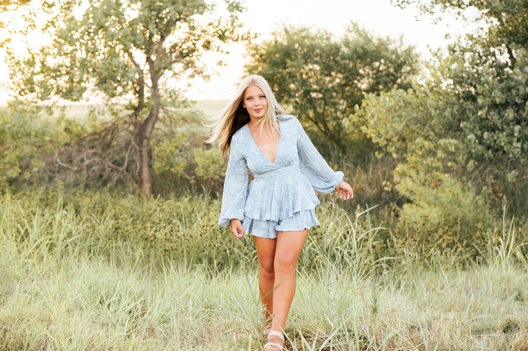 High school senior girl walks toward the camera in a blue dress with her arms out at her sides in a field in central Oklahoma for her senior photos