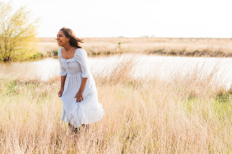 A high school senior girl laughs while she stands in a golden field with a pond for a senior portrait session in Oklahoma