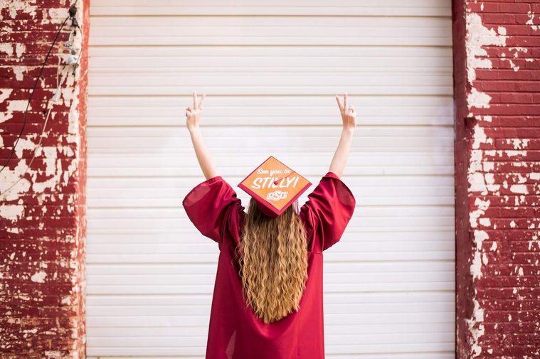 high school senior girl with long blond hair wearing her red cap and graduation gown faces away from camera with both hands as peace signs with a college sticker on top of her cap in front of a rustic garage door in midtown Oklahoma City
